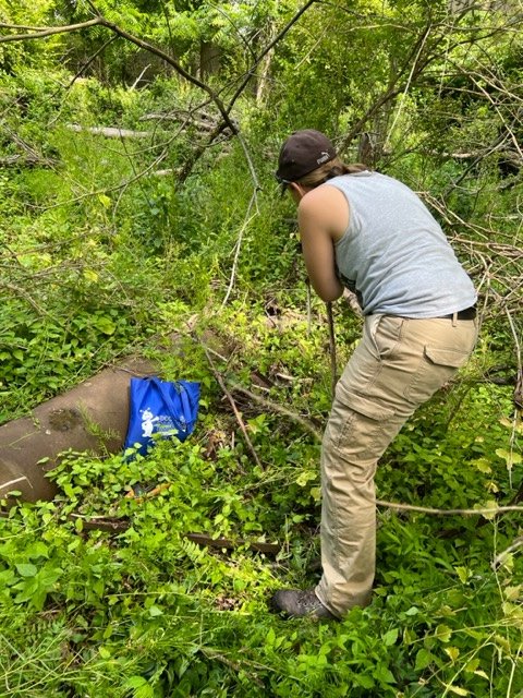 A volunteer bent over, carefully digging near a buried object, with a tool bag beside them.